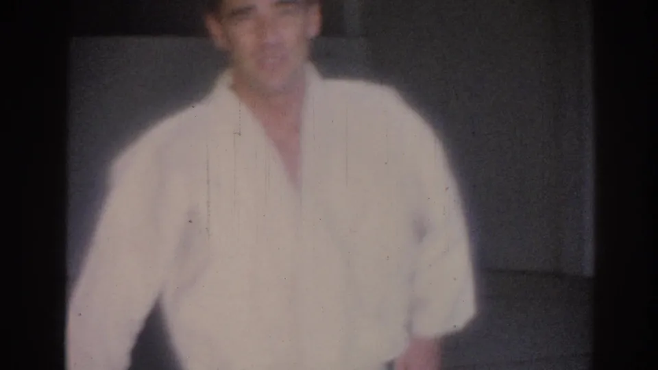 1967 Los Angeles home-movie frame showing a young man in a white judo gi facing the camera on an indoor mat.