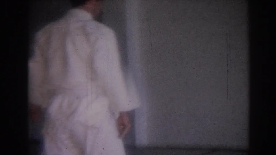 1967 Los Angeles home-movie frame showing a martial arts practitioner in a white gi from behind in a simple gym space.