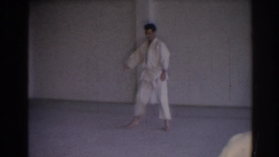 1967 Los Angeles frame showing a martial arts practitioner in a white gi standing in a gym, with a partial foreground observer.