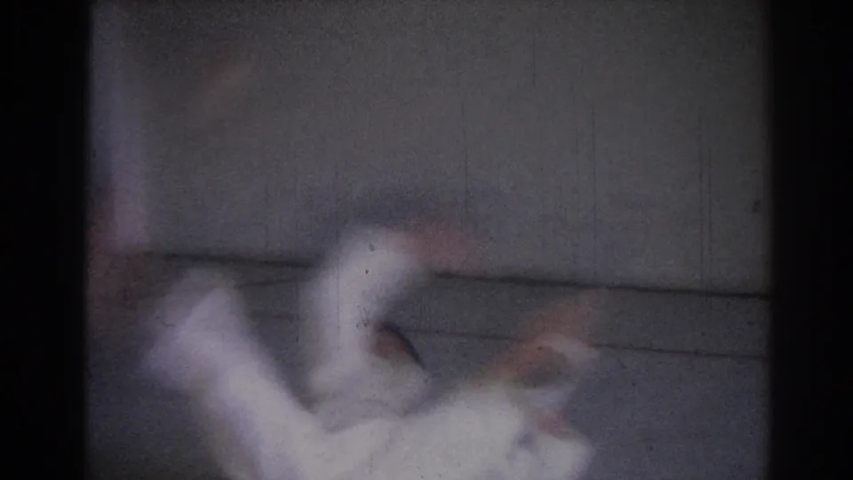 A soft 8mm frame shows the white-gi practitioner in motion during a fall or roll on the gym mat.