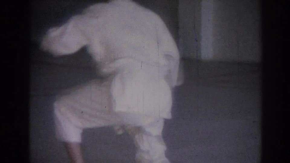1967 Los Angeles 8mm frame showing a man in a white karate uniform crouched low inside a sparse room.