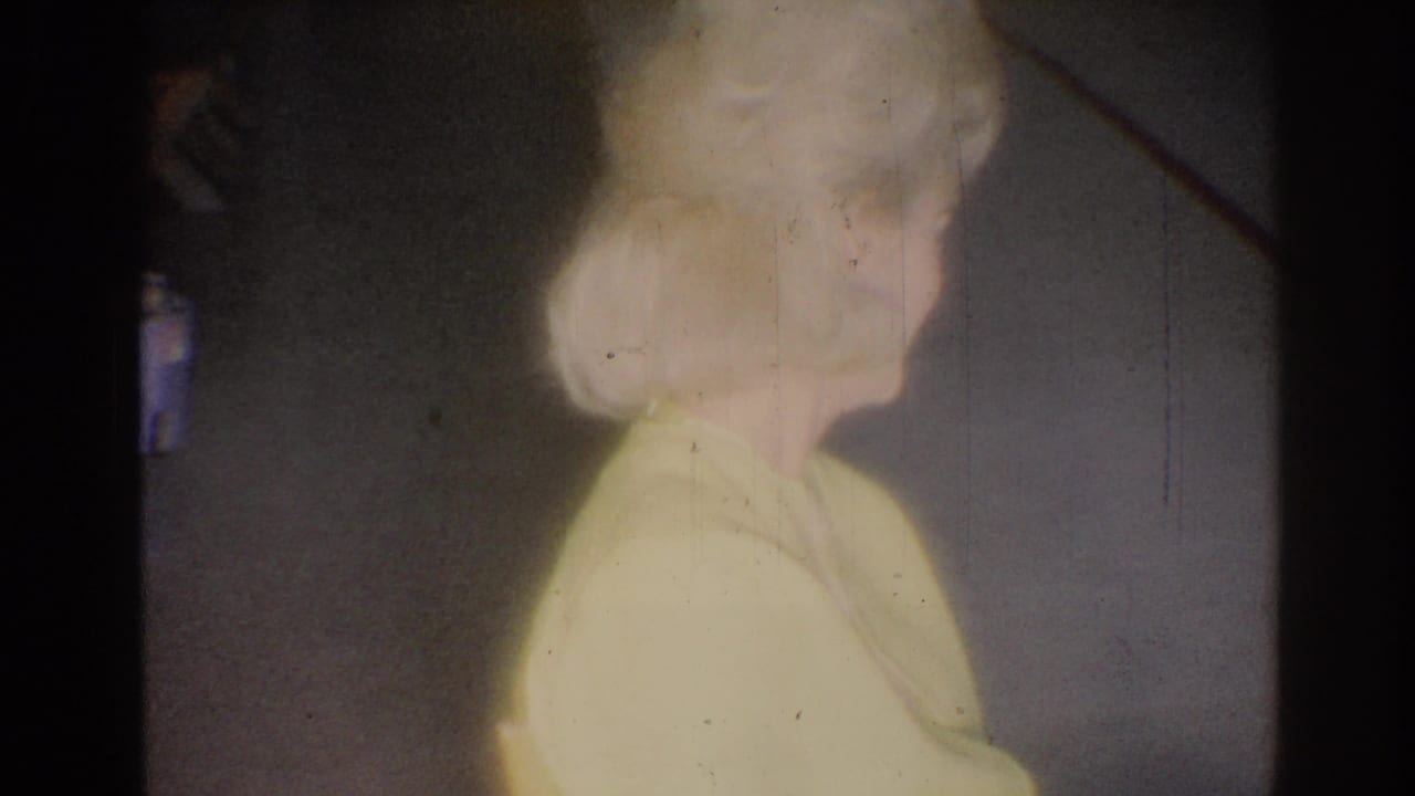 Blonde woman in a yellow blouse smiles while looking to the side indoors.
