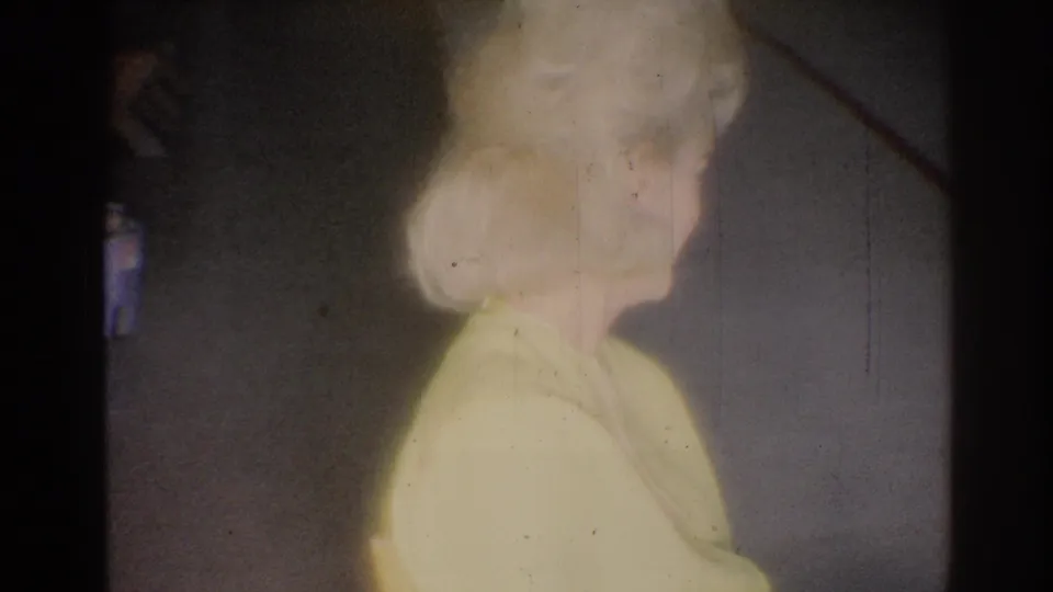 1967 Los Angeles 8mm frame of a blonde woman in a yellow blouse watching from the side in a plain indoor setting.