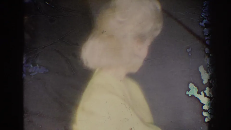 1967 Los Angeles 8mm frame showing the woman in yellow from behind and side as she watches inside a simple room.