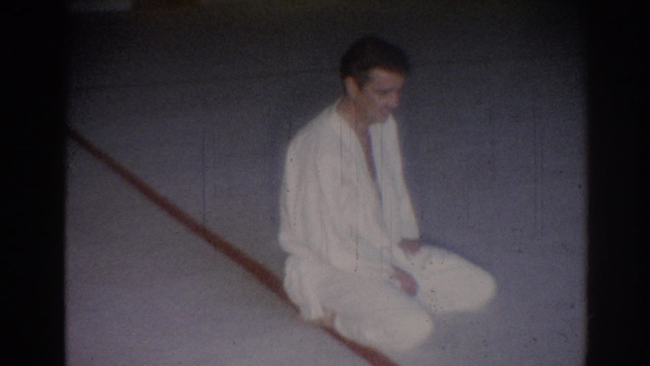 Young man in a white gi seated on a gray floor near a red line.