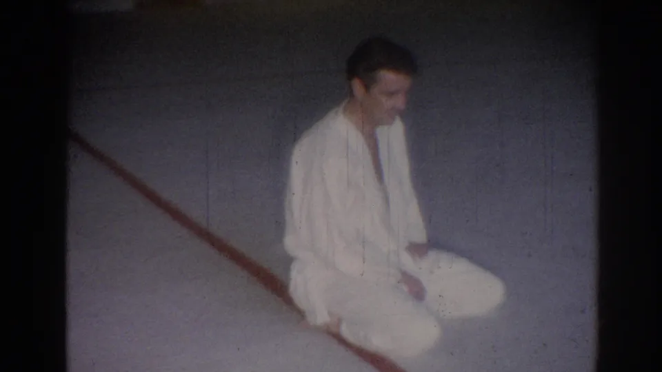 Young man in a white martial arts gi sits on a gray gym floor near a red line in this 1967 Los Angeles home-movie frame.