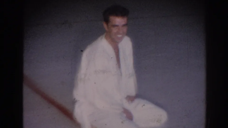 In a soft 8mm frame, the seated young man in a white gi smiles toward the camera on a gray floor with a red line.