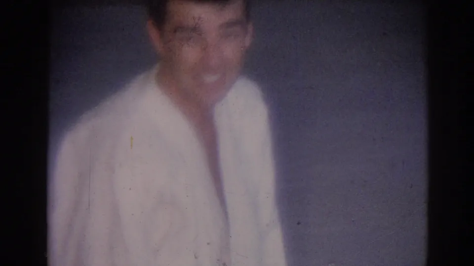 Soft close view of a smiling young man in a white gi, with grain, fading, and scratches visible in the 8mm scan.