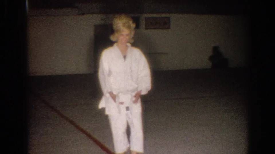 The woman in the white gi looks down while standing in the dim Los Angeles practice room recorded on 8mm film.