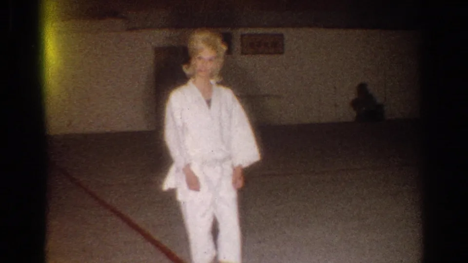 A woman in a white gi faces the camera, with a red floor line and shadowed wall visible in the 1967 home-movie frame.