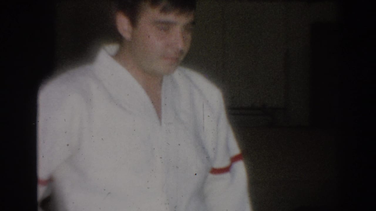 Young man in a white judogi with red sleeve trim seated or standing in a dark room.