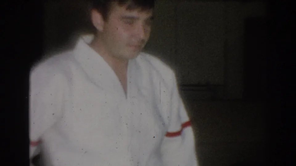 1967 Los Angeles 8mm frame with a young judo participant in a white judogi with red sleeve trim, softly lit in an interior space.