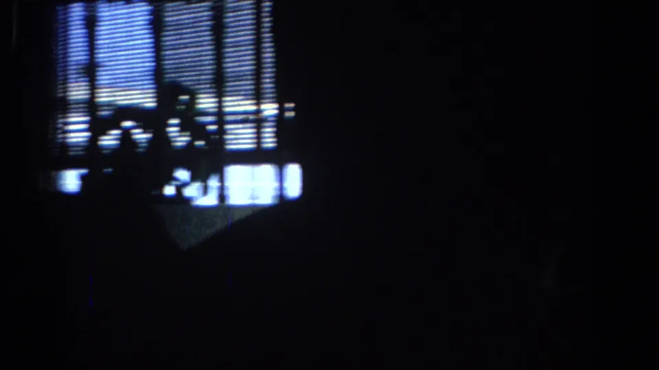 Dim 1967 Los Angeles home-movie frame showing window blinds, strong backlight, and silhouetted people inside the competition space.