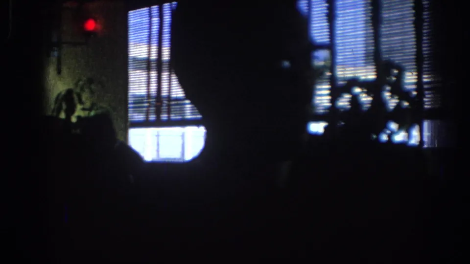 1967 Los Angeles 8mm frame of a dim interior with window blinds, silhouetted people, and a small red light in the background.