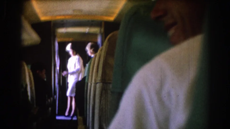 Seat-level view inside a 1967 Los Angeles train interior, with upholstered seats, a doorway, and passengers including a woman in a light suit and hat.