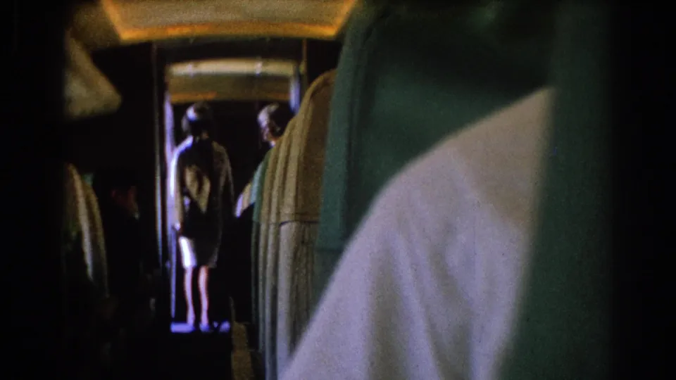 Passenger-seat view inside the 1967 Los Angeles train clip, showing a dim aisle, upholstered seats, and figures standing in the doorway area.