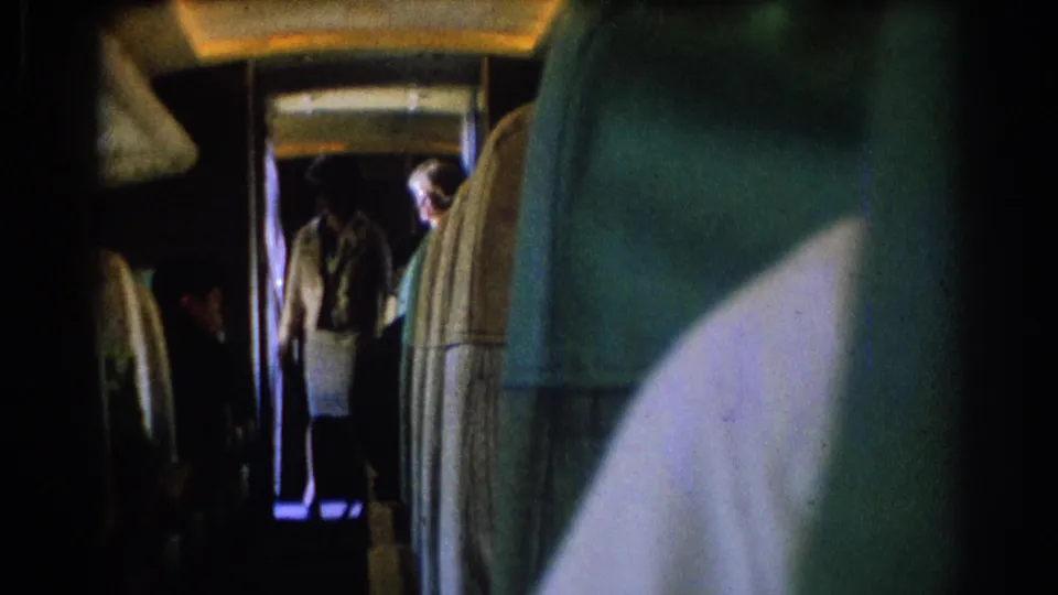 1967 8mm airplane cabin view from a seat, showing a narrow aisle, upholstered seats, passengers, and a flight attendant-like figure in the background.