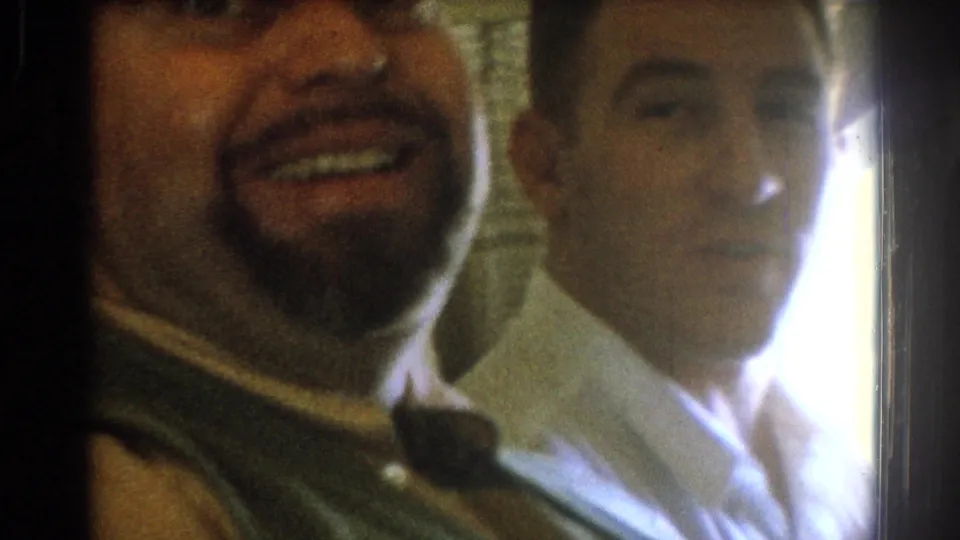 Close 1967 airplane-cabin shot of two adult passengers, with the foreground man smiling toward the camera and a second passenger lit from the side.