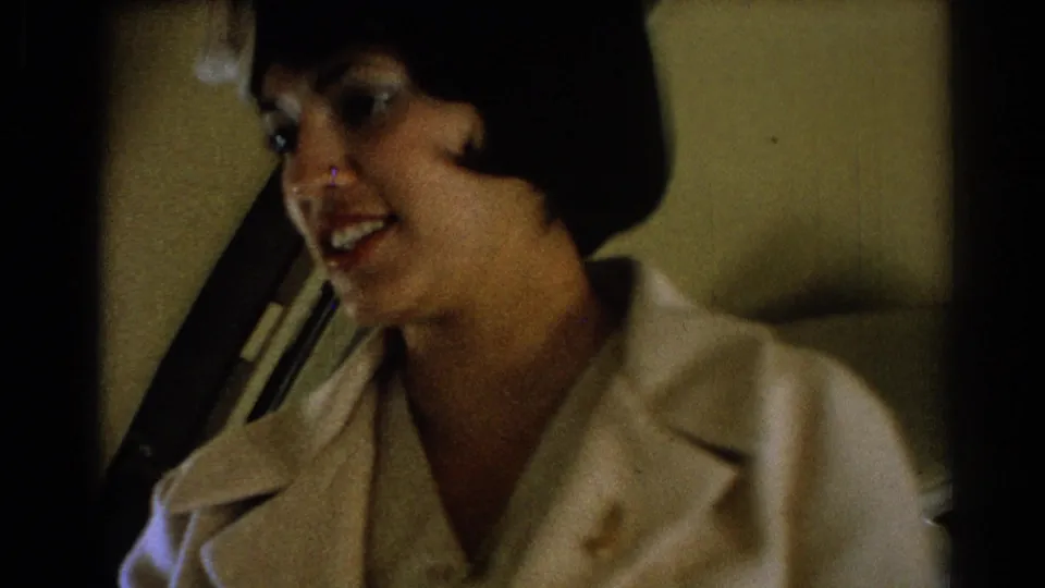Woman in a light flight-attendant-style uniform appears beside an airplane seat, smiling in a softly lit 1967 8mm cabin view.