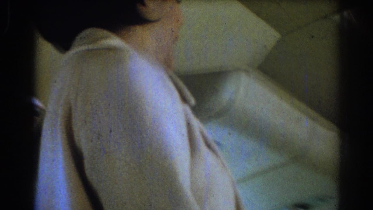 The flight attendant turns to the side in the aircraft cabin, with her light uniform and the patterned seat upholstery visible in soft, grainy 8mm footage.