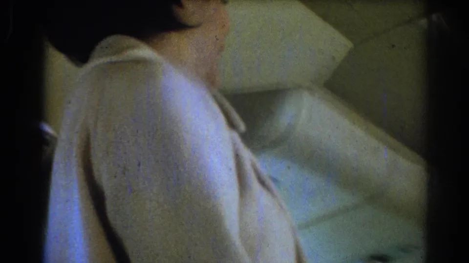 The flight attendant turns to the side in the aircraft cabin, with her light uniform and the patterned seat upholstery visible in soft, grainy 8mm footage.