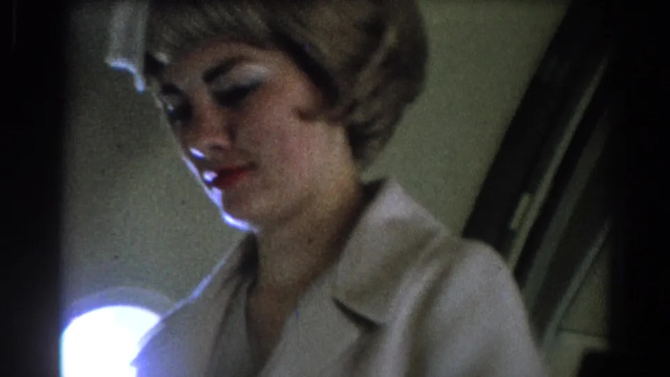 A stewardess with a 1960s hairstyle stands in a dim aircraft cabin, partially lit from below and behind.