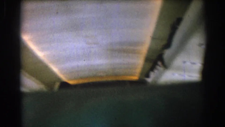 Low-angle view of airplane cabin ceiling panels with warm overhead lighting in grainy 8mm footage.