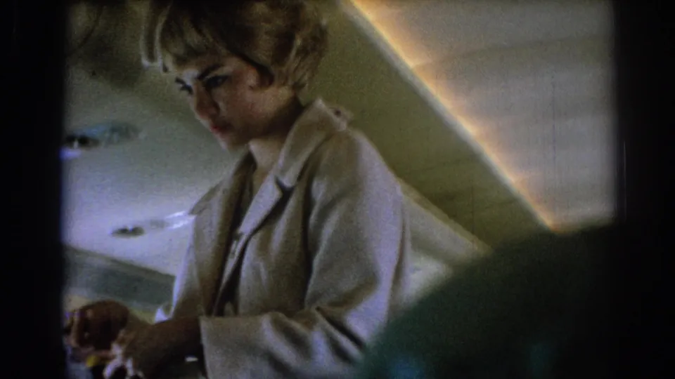 Wider passenger-view frame of a stewardess in a light uniform beneath softly lit 1967 airplane cabin panels.
