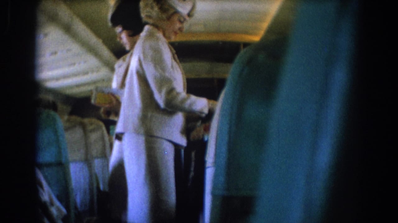 Two uniformed flight attendants stand in an airplane aisle near passenger seats.