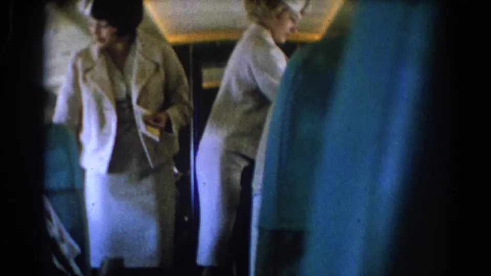A female flight attendant holds an item in the aisle while another attendant stands farther back in the airplane cabin.