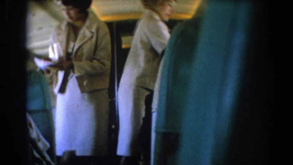 Inside the airplane cabin, one uniformed attendant stands near center frame while another moves along the seats.