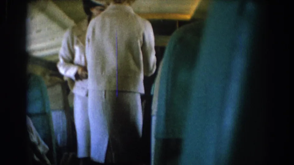 A white-uniformed flight attendant is seen from behind in a 1967 airplane cabin, with another attendant partly visible ahead.