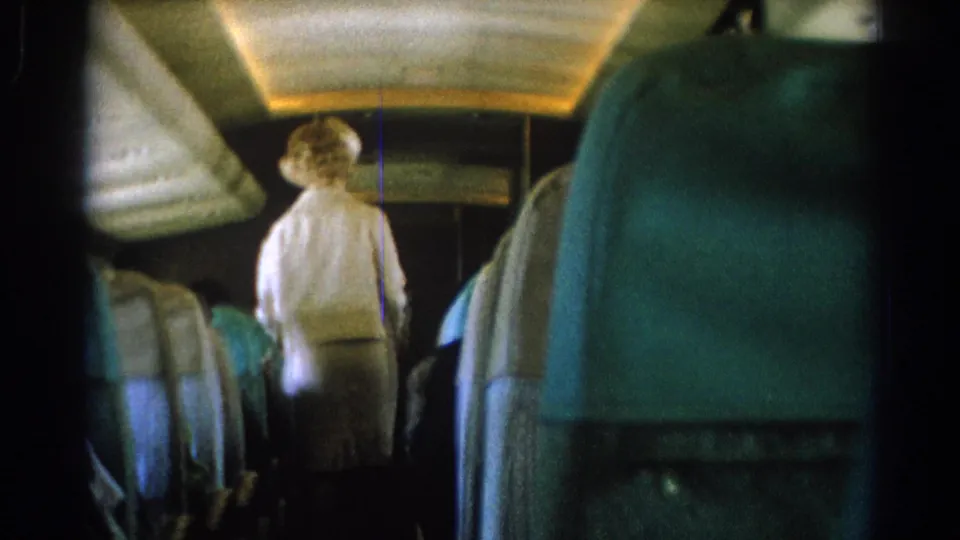 From a passenger seat, the 1967 8mm frame shows a flight attendant walking down the airplane aisle between teal-blue rows.