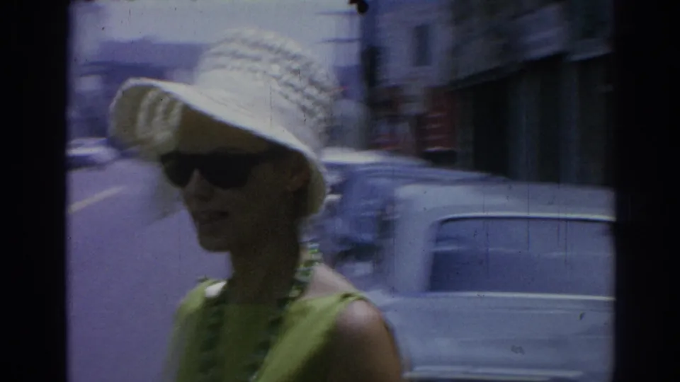 In 1967 Los Angeles street footage, a woman in a white straw hat, dark sunglasses, green dress, and necklace walks past blurred cars.
