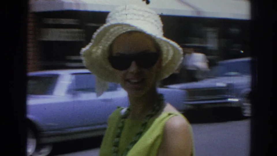 A woman in a white straw hat, sunglasses, green dress, and necklace smiles toward the camera on a Los Angeles street in 1967 footage.
