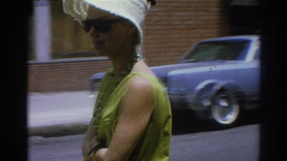 Adult woman in 1967 Los Angeles wearing a lime green sleeveless dress, white sunhat, and dark sunglasses as she walks along a sidewalk with a blue car behind her.