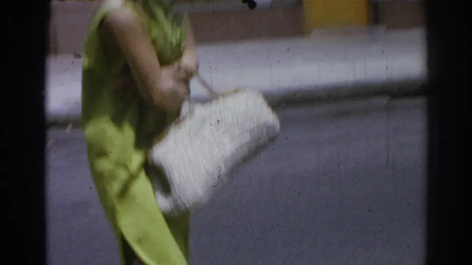 Close side view of an adult woman in a lime green dress carrying a large white woven purse while walking on a Los Angeles sidewalk.