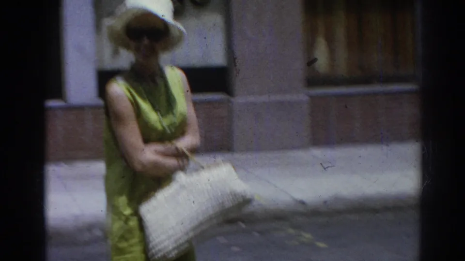 Adult woman in white sunhat, sunglasses, and lime green dress stands or walks near a building facade while holding a large white woven purse.
