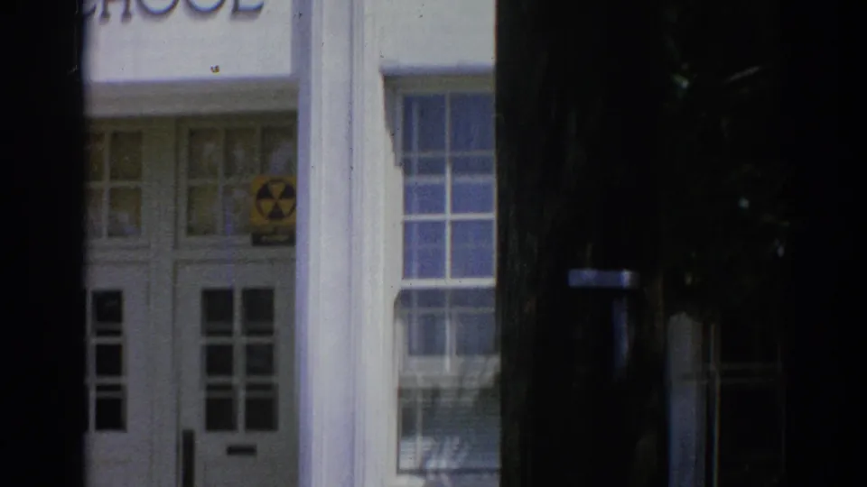 1967 Los Angeles 8mm frame showing a white institutional entrance with columns, multi-pane doors, steps, and a yellow radiation warning symbol.