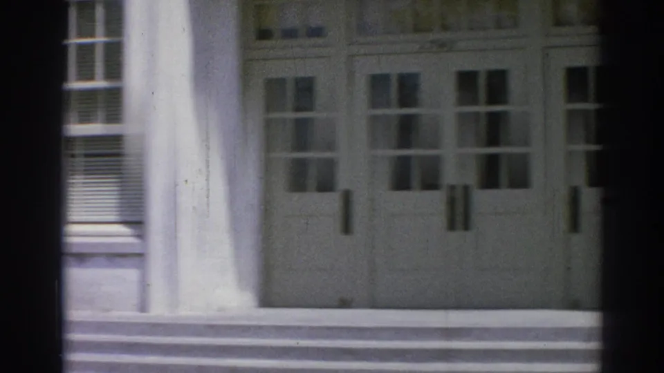 1967 Los Angeles 8mm frame of a white institutional facade with double doors, column bases, steps, and multi-pane windows.