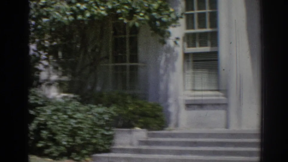 1967 Los Angeles 8mm frame showing steps and windows on a white institutional building partly shaded by trees and shrubs.