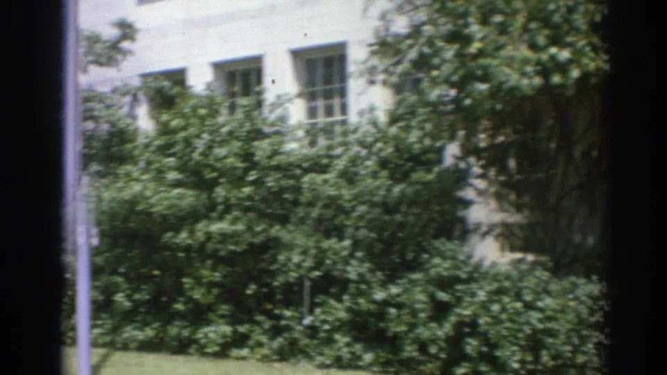 1967 Los Angeles 8mm frame of a landscaped institutional exterior with white walls, windows, dense shrubs, lawn, and tree branches.