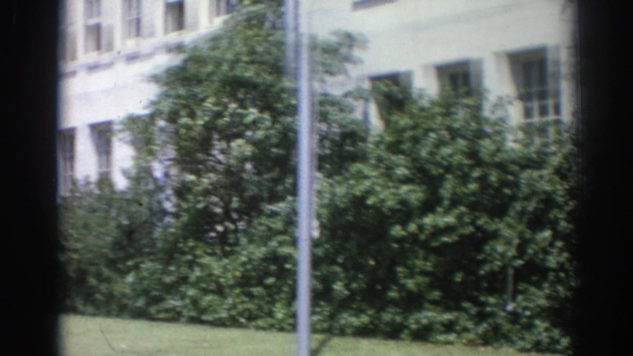 Pale institutional building behind hedges and a lawn, framed by dark vertical edges.