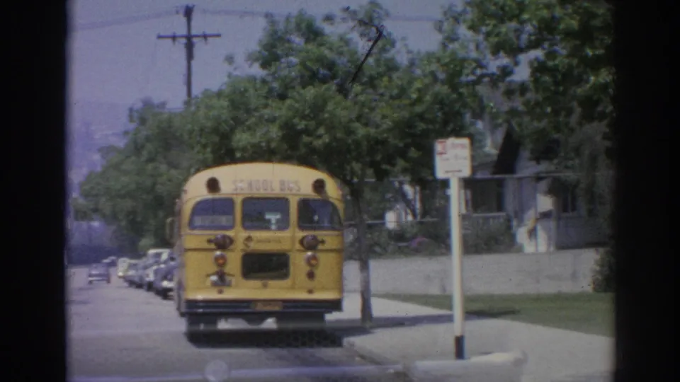 1967 Los Angeles street view with a yellow school bus, parked cars, trees, houses, and a roadside sign.