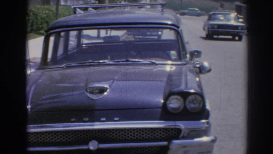 Close 1967 Los Angeles street view of a dark Ford station wagon with roof rack and another car in the lane behind.