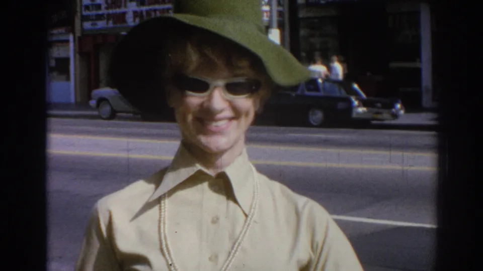 Woman in a wide green hat, white sunglasses, light collared shirt, and pearl necklace smiles on a Los Angeles street in 1967 footage.