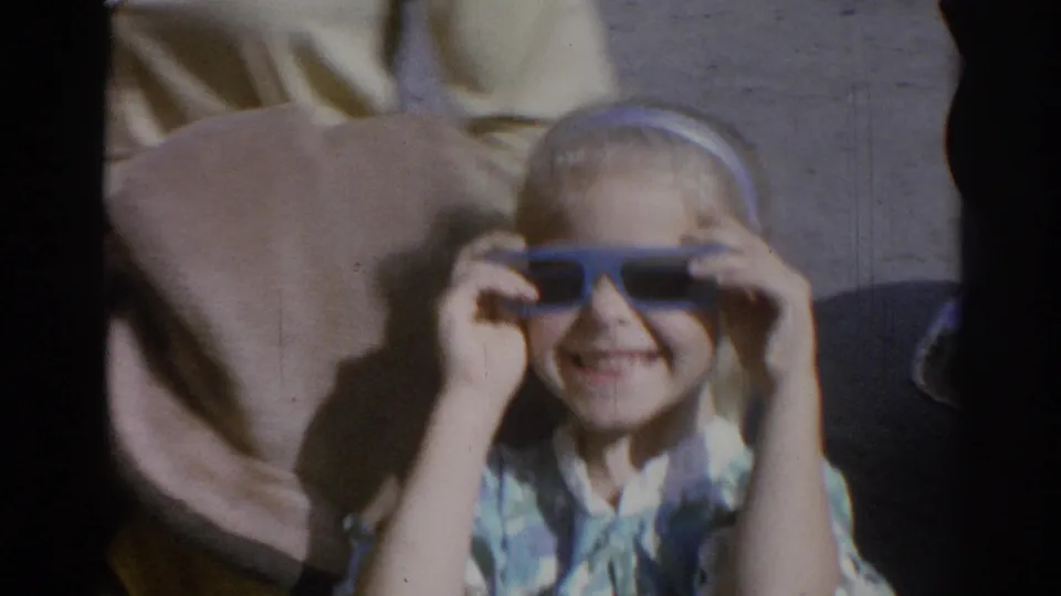 Child in a car seat holds dark sunglasses while an adult in a light shirt is partly visible beside them.