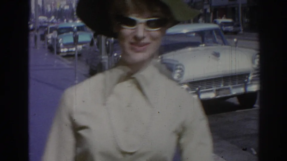 Woman in green hat and white sunglasses stands beside a street lined with parked mid-century cars in 1967 Los Angeles footage.