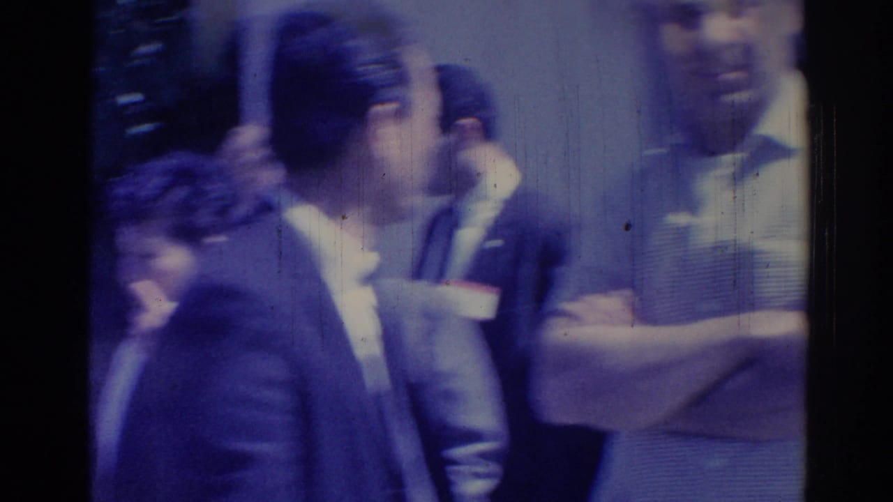 Adults in suits and name tags stand in a crowded indoor or shaded event area.