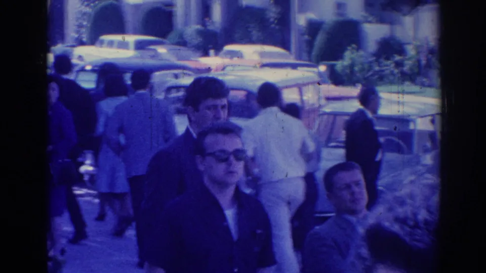 1967 Los Angeles home-movie frame showing adults walking outside near parked mid-century cars in bright daylight.